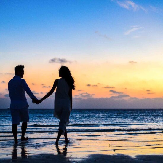 man and woman holding hands walking on seashore during sunrise